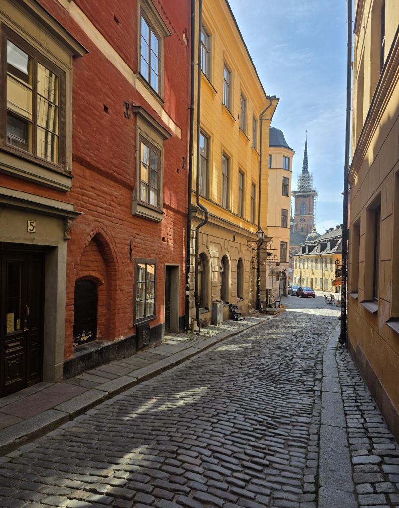Kopfsteinpflastergasse mit roten und gelben Häusern und Kirchturm im Hintergrund in Gamla Stan.
