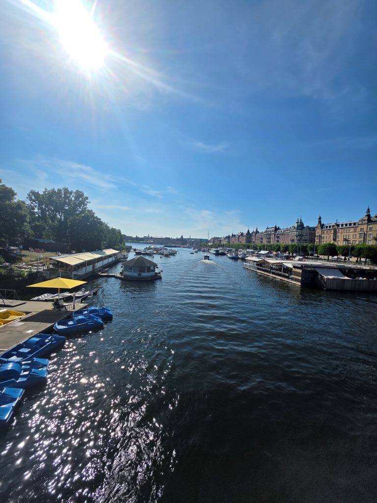 Blick über das Wasser in Stockholm mit Booten, Uferpromenade und historischen Häusern bei Sonnenschein.