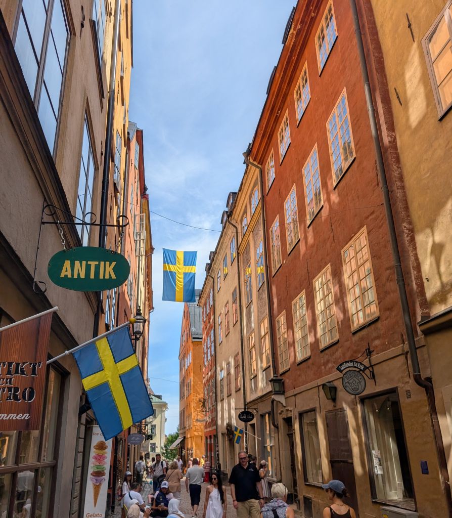 Belebte Gasse in Gamla Stan mit bunten Häusern, schwedischen Flaggen und kleinen Geschäften.