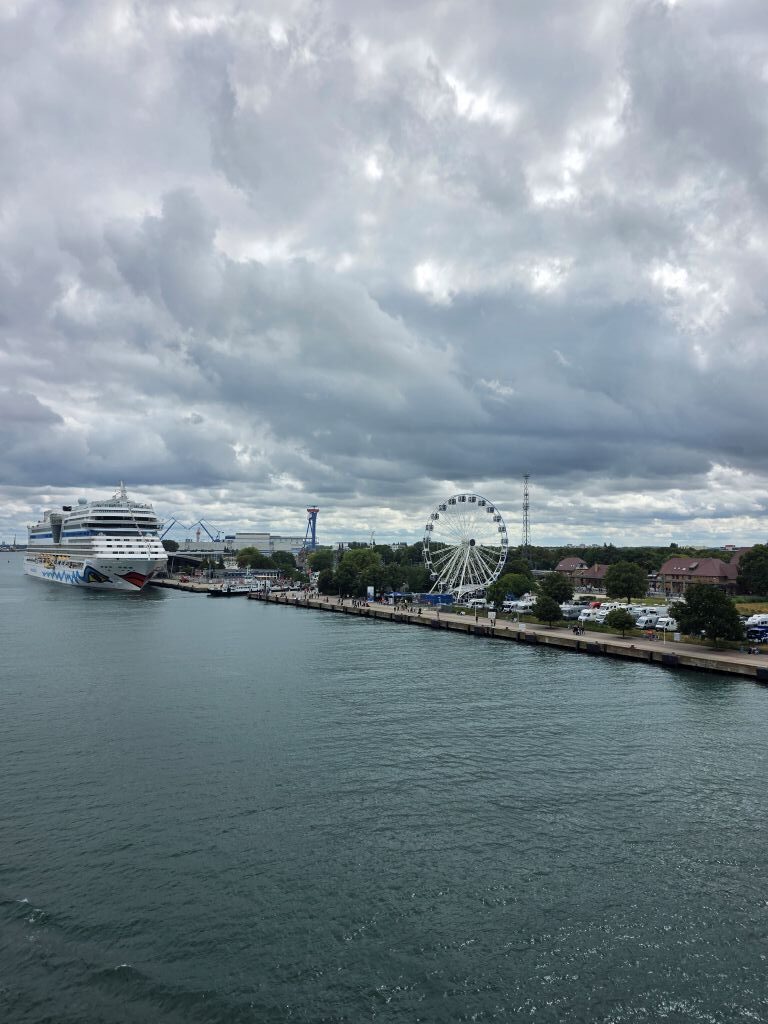 Hafeneinfahrt in Rostock mit Kreuzfahrtschiff und Riesenrad unter dunklen Wolken.