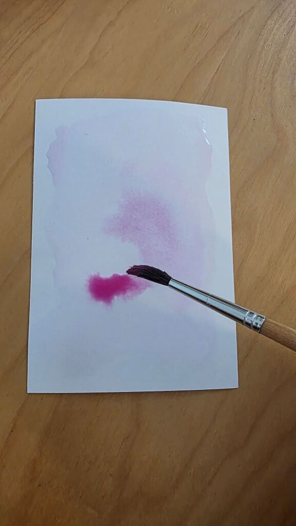 Rosa Wasserfarbe wird mit einem Pinsel auf den weißen Karton der Konfirmationskarte aufgetragen.