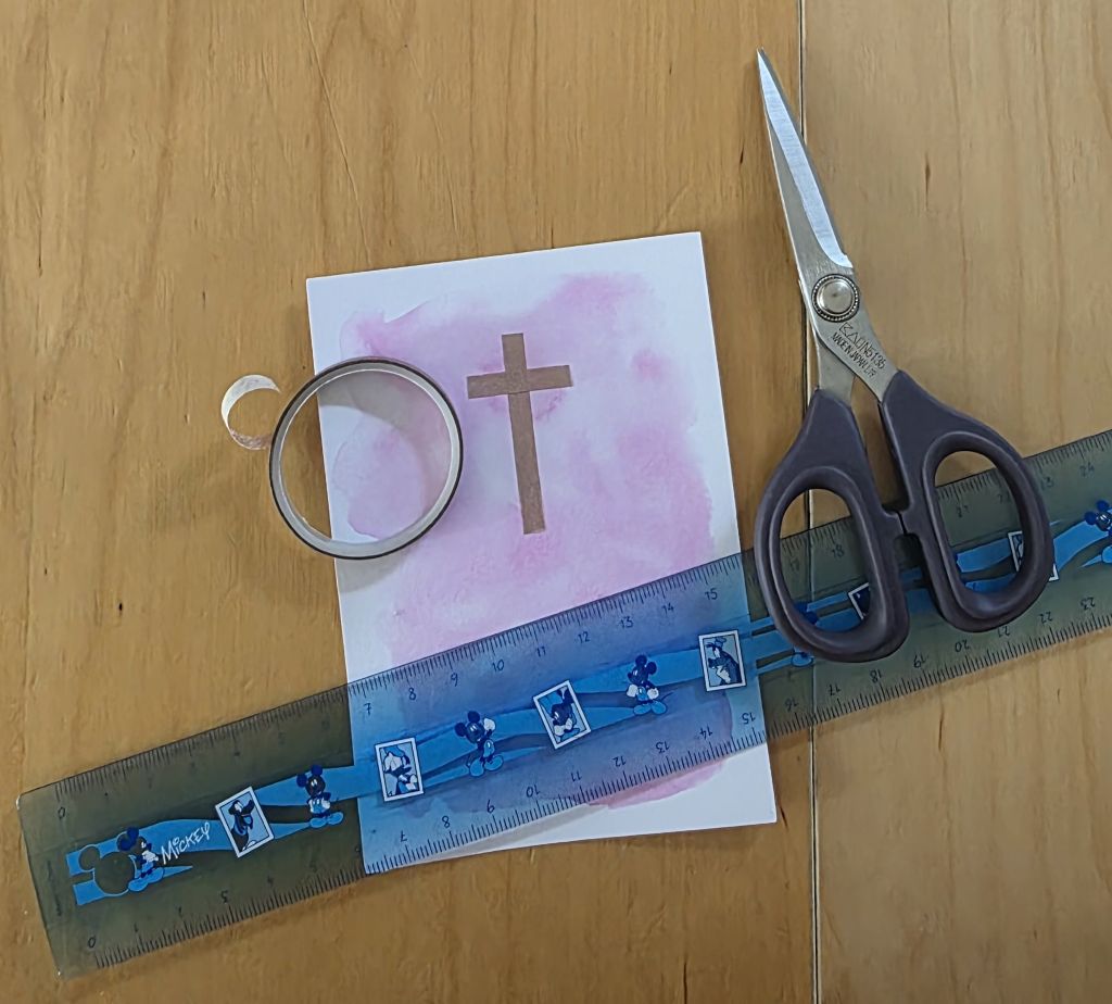 Weißer Karton mit rosa Aquarellhintergrund und Kreuz aus Washi Tape, daneben Schere, Lineal und Washi Tape.