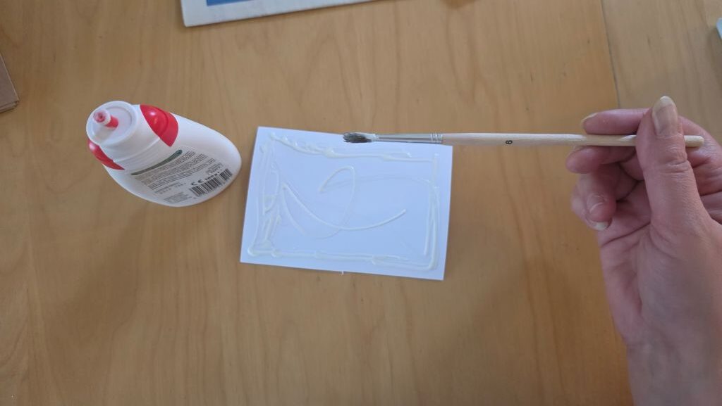 Bastelkleber wird mit einem Pinsel auf die Rückseite des weißen Kartons für die Konfirmationskarte verteilt.